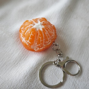 May include: A novelty keychain featuring a detailed, orange mandarin orange segment design. The keychain has a silver-coloured metal clasp and ring. The background is a white fabric.