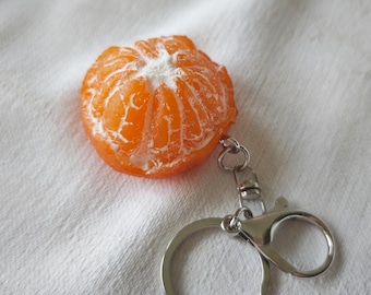 May include: A realistic orange keychain shaped like a peeled mandarin orange. The keychain is bright orange with white segments and a silver metal clasp and ring. The keychain is on a white fabric background.