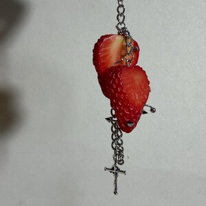 May include: A silver-toned metal keychain featuring two realistic red strawberry slices attached to a link chain. The accessory includes a small silver cross charm and a metal spike stud detail, all suspended from a lobster claw clasp.
