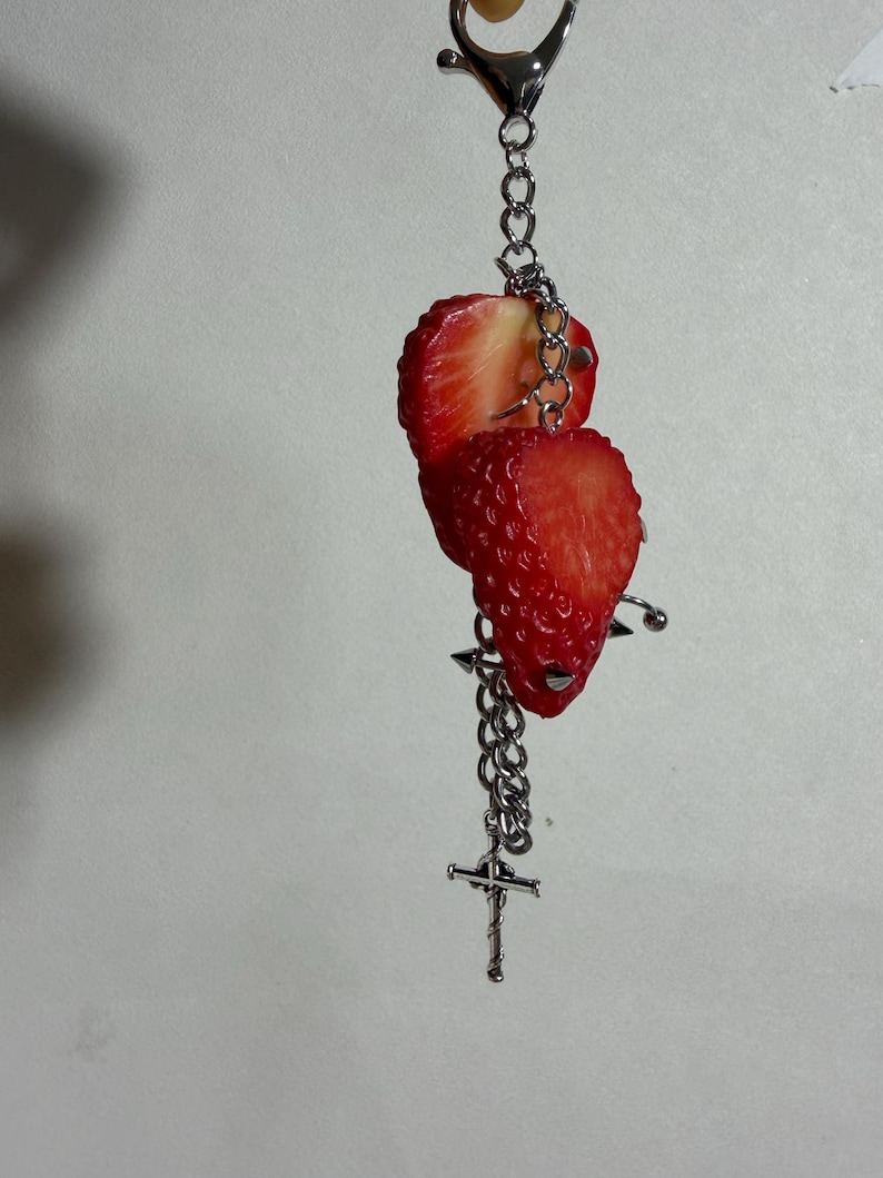 May include: A silver-toned metal keychain featuring two realistic red strawberry slices attached to a link chain. The accessory includes a small silver cross charm and a metal spike stud detail, all suspended from a lobster claw clasp.