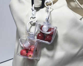 May include: Two clear, square, plastic containers filled with red apples, attached to a white clip. The containers are hanging from a white bag. The apples are a deep red color.