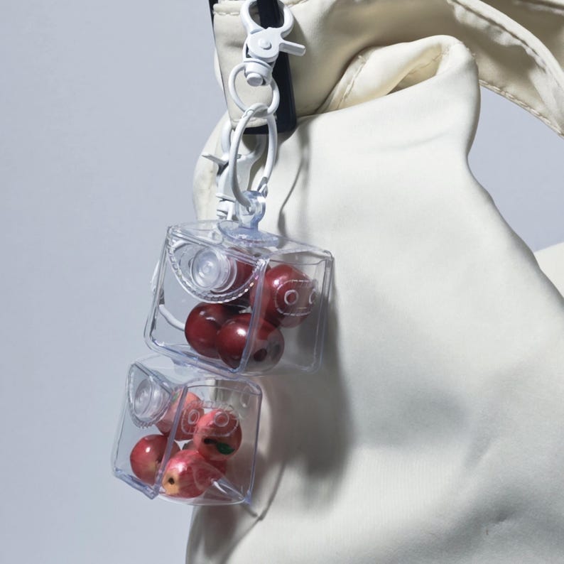 May include: Two clear, cube-shaped containers, each filled with red, round objects, hang from a white clip. The cubes are attached to a white strap, set against a soft, off-white fabric background. The objects inside the cubes have small, painted faces.
