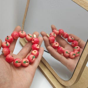 May include: A bracelet featuring a series of small, red apple charms with green leaves. The bracelet is held in a hand, with a mirror reflecting the bracelet and hand. The apples have a realistic appearance.