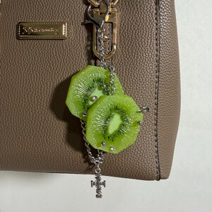 May include: A taupe textured handbag with a silver-toned metal keychain attached to the handle. The keychain features two realistic green kiwi fruit slices adorned with small silver studs and a dangling silver cross-shaped charm on a chain.