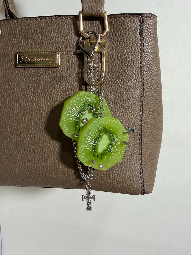 May include: A taupe textured handbag with a silver-toned metal keychain attached to the handle. The keychain features two realistic green kiwi fruit slices adorned with small silver studs and a dangling silver cross-shaped charm on a chain.