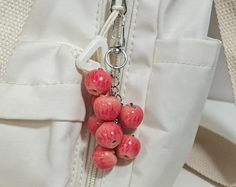 May include: A white fabric bag featuring a silver-toned metal keychain attachment. The keychain consists of a cluster of small, glossy, red apple-shaped charms suspended from a silver chain and a white triangular plastic connector piece.