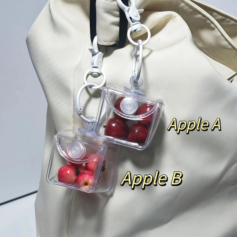 May include: Two clear, square, plastic containers, each filled with red apples, are suspended from white clips. The text "Apple A" and "Apple B" are printed in yellow. The background is a cream-coloured fabric.