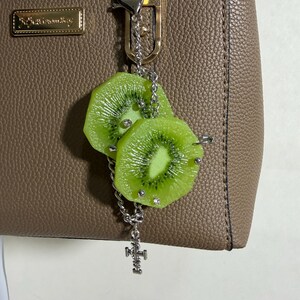 May include: A silver-toned metal keychain featuring two realistic green kiwi fruit slices attached to a link chain. The kiwi slices are accented with small silver studs and clear rhinestones. A small silver cross charm hangs from the bottom of the chain.