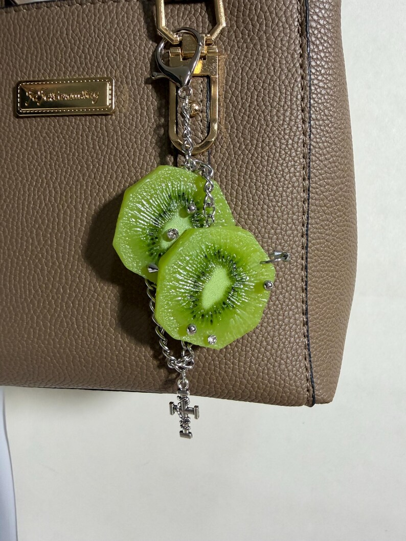 May include: A silver-toned metal keychain featuring two realistic green kiwi fruit slices attached to a link chain. The kiwi slices are accented with small silver studs and clear rhinestones. A small silver cross charm hangs from the bottom of the chain.