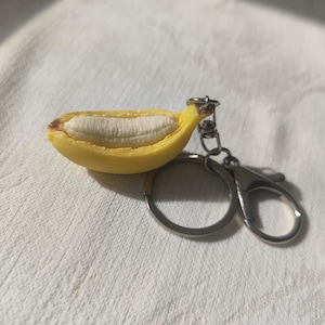 May include: A yellow banana keyring with a peeled design, showing the white fruit within. The keyring has a silver-coloured ring and clasp. The banana peel has brown accents at the ends.
