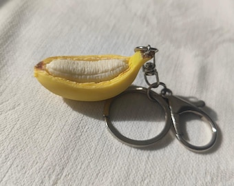 May include: A yellow banana keychain with a peeled design, revealing the white fruit inside. The keychain has a silver-colored ring and clasp. The banana peel has brown accents at the ends.