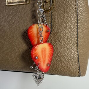 May include: A silver-toned metal chain bag charm featuring two sliced strawberry charms with small silver spikes, a small clear crystal bead, and a dangling heart-shaped pendant with engraved text, attached to a textured taupe handbag.