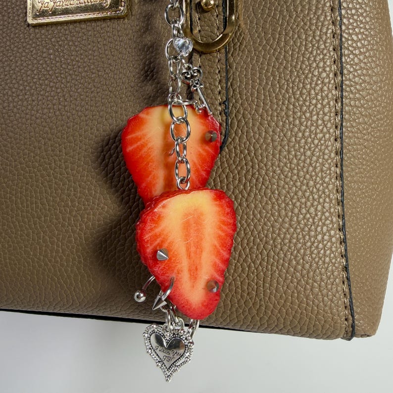 May include: A silver-toned metal chain bag charm featuring two sliced strawberry charms with small silver spikes, a small clear crystal bead, and a dangling heart-shaped pendant with engraved text, attached to a textured taupe handbag.