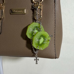 May include: A taupe textured handbag with gold-tone hardware and a rectangular logo plate. Attached to the handle is a silver-tone chain charm featuring two realistic green kiwi fruit slices adorned with small silver studs and a small cross pendant.