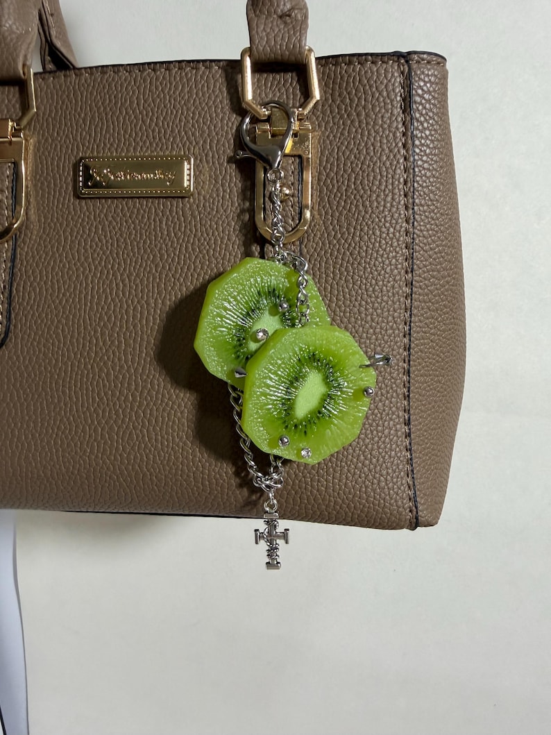May include: A taupe textured handbag with gold-tone hardware and a rectangular logo plate. Attached to the handle is a silver-tone chain charm featuring two realistic green kiwi fruit slices adorned with small silver studs and a small cross pendant.