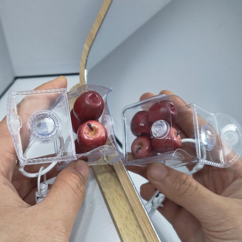 May include: Two clear plastic containers, each holding several small, red artificial apples. The containers have suction cups and white cords. The background includes a wooden element and a white surface.