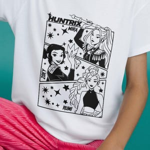 May include: White t-shirt featuring a black and white graphic of three women in a comic book style. The text "HUNTRIX" and the names "MIRA", "ZOEY", and "RUMI" are included. The shirt is worn by a person wearing pink pants.