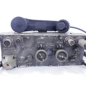 May include: A vintage military radio transceiver in olive drab metal, with a black handset. The unit has various dials, connectors, and labels, including "ANT" and "ALIMENTAÇÃO". A coiled cable extends from the side.