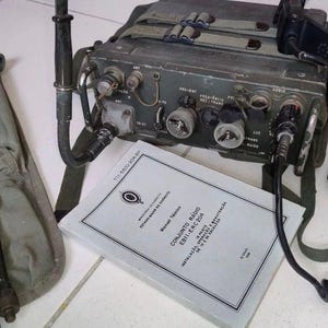 Könnte beinhalten: Ein olivgrünes militärisches Funkgerät mit einem spiralförmigen schwarzen Höreranschlusskabel, einer vertikalen Antenne und einem Trageriemen. Daneben liegt eine olivgrüne Antennentasche aus Segeltuch mit Metallstäben und einem technischen Handbuch.