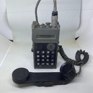 May include: A dark grey portable military radio transceiver featuring a rectangular display screen, a black keypad with alphanumeric buttons, and a connected black handset with a coiled cord. The device has a top-mounted antenna port and cable.