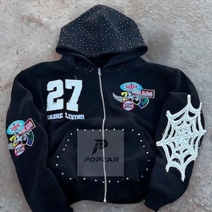 May include: Black zip-up hoodie with a studded hood and pocket trim. Features the number "27" and patches with text and graphics. A white spiderweb design is on the sleeve. The hoodie has a silver zipper.