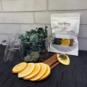 May include: A clear glass jar with a lid, filled with green eucalyptus leaves, sits next to a white pouch labeled "Stress Relief." Dried orange slices and cinnamon sticks are arranged near the jar.