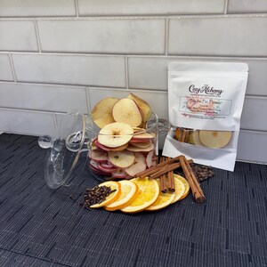 May include: A clear glass jar filled with dried apple slices, next to a white pouch with the text "Cozy Alchemy". Also, orange slices, cinnamon sticks, cloves, and peppercorns are arranged on a dark surface.