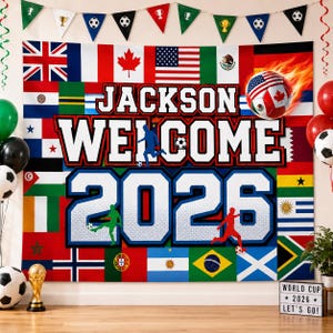May include: A rectangular fabric backdrop featuring a collage of international flags, the text "JACKSON WELCOME 2026," and silhouettes of soccer players. A flaming soccer ball graphic is positioned in the upper right corner of the banner.