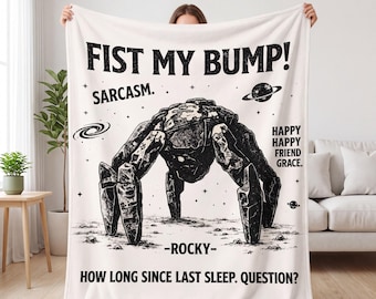 May include: A white fleece blanket featuring a black graphic of a rock creature. Text reads: FIST MY BUMP! SARCASM. HAPPY HAPPY FRIEND GRACE. -ROCKY- HOW LONG SINCE LAST SLEEP. QUESTION? The design includes small stars, a planet, and a spiral galaxy.