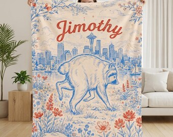 May include: A cream-colored throw blanket featuring a blue line-art illustration of a raccoon walking through a field of red and blue flowers. In the background, a blue city skyline includes the Space Needle. The name Jimothy is printed in red.