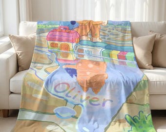 May include: A soft fleece blanket featuring a colorful cartoon illustration of two dog characters standing near a puddle. The puddle reflects the characters and a small toy house. The name Oliver is printed in large, light-colored letters.