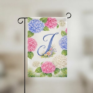 May include: A garden flag featuring a watercolor design with a large blue letter "J" surrounded by pink, blue, and white hydrangea flowers. The flag is displayed on a black metal stand.