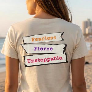 May include: A beige t-shirt with the words "Fearless," "Fierce," and "Unstoppable" printed on the back in orange, purple, and red. The shirt is paired with denim shorts. The image is taken on a beach with a boardwalk and ocean in the background.