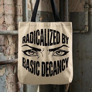 Może przedstawiać: Beżowa torba typu tote z czarnymi uchwytami, zawieszona na metalowym haczyku. Torba zawiera napis "RADICALIZED BY BASIC DECENCY" w kolorze czarnym, ze stylizowanymi oczami poniżej.