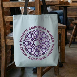 May include: A light blue tote bag with black handles, hanging on a wooden chair. The bag features a purple circular design with the words "EMPOWER WOMEN" around the edge. The bag is in a cafe setting.