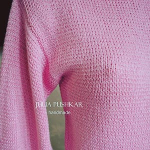 May include: A pale pink, handmade knitted jumper with long sleeves. The jumper features a boat neck and is crafted from a soft, textured yarn. The text "JULIA PUSHKAR handmade" is visible on the front of the jumper.