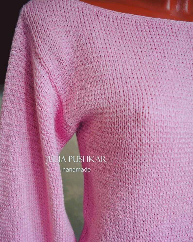 May include: A pale pink, handmade knitted jumper with long sleeves. The jumper features a boat neck and is crafted from a soft, textured yarn. The text "JULIA PUSHKAR handmade" is visible on the front of the jumper.