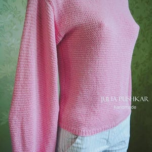 May include: A pale pink knitted sweater with long sleeves and a boat neck. The sweater is made from a textured knit fabric. The sleeves are long and full. The text "julia pushkar handmade" is visible on the right side of the sweater.