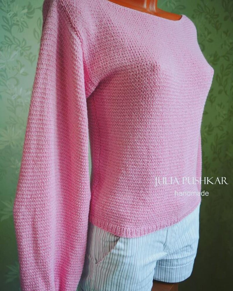 May include: A pale pink knitted sweater with long sleeves and a boat neck. The sweater is made from a textured knit fabric. The sleeves are long and full. The text "julia pushkar handmade" is visible on the right side of the sweater.