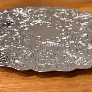 May include: A silver-colored, ornate plate with a scalloped edge. The plate features a detailed, embossed floral and scroll design. The plate is round and sits on a wooden surface. The plate is likely used for serving or display.