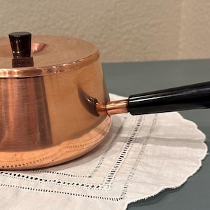 May include: A copper saucepan with a black handle and lid. The small pot sits on a white, scalloped-edge doily. The handle is angled and attached to the side of the pot. The lid has a small, black knob.