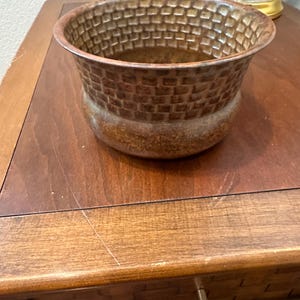 May include: A brown ceramic planter with a basket weave design. The planter has a wide, open top and a rounded base. The color is a warm, earthy brown, and it sits on a wooden surface.