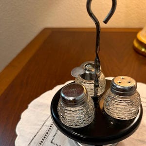 Op de afbeelding: Een vintage zout- en peperstrooier set met een suikerdispenser. De set heeft drie transparante glazen containers met getextureerde zijkanten en zilverkleurige metalen deksels. De containers staan op een zwarte cirkelvormige basis met een metalen handvat.