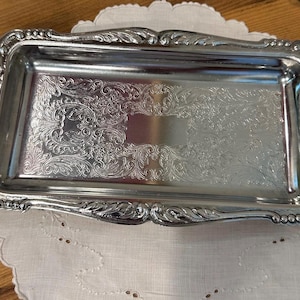 May include: A rectangular silver-toned metal tray featuring an ornate embossed border with shell-like motifs at the ends and intricate scrollwork patterns engraved across the central flat surface, creating a reflective and polished finish.