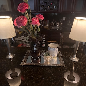 May include: Two silver table lamps with off-white shades flank a mirrored tray with a vase of pink flowers, a candle, and a small glass cup. The lamps have a classic, elegant design, perfect for home decor.