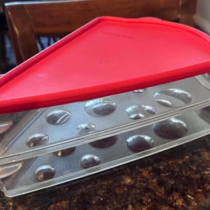 May include: A triangular, clear plastic food storage container with a bright red lid. The container features two stacked, transparent sections with a textured, dimpled surface pattern on the sides, resembling a slice of Swiss cheese.
