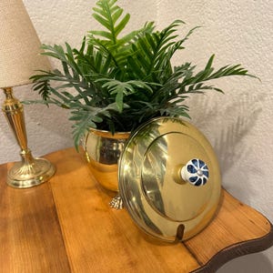 May include: A gold-colored metal container with a lid featuring a white and blue floral design knob. The container sits on a wooden table next to a gold lamp and a potted green plant. The container is round and has small decorative feet.