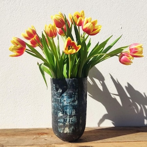 Könnte beinhalten: Ein Strauß gelb-roter Tulpen in einer hohen, strukturierten, dunkelblauen Vase. Die Vase steht auf einer Holzoberfläche vor einer weißen Wand. Das Sonnenlicht wirft einen Schatten der Blumen.
