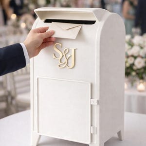 Op de afbeelding: Een witte trouwkaartendoos met een gleuf voor kaarten en een scharnierende deur. De doos heeft gouden initialen "S&J" op de voorkant. Een hand steekt een kaart in de gleuf. De doos staat op een witte tafel.