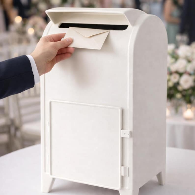 Op de afbeelding: Een witte brievenbus met een gewelfde bovenkant en een gleuf voor het plaatsen van post. De brievenbus heeft een deur aan de voorkant en staat op vier kleine poten. Een hand steekt een cr&egrave;mekleurige envelop in de gleuf.